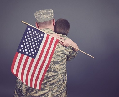 Back of US military soldier with the United States flag showing over his shoulder and back. He is hugging a child